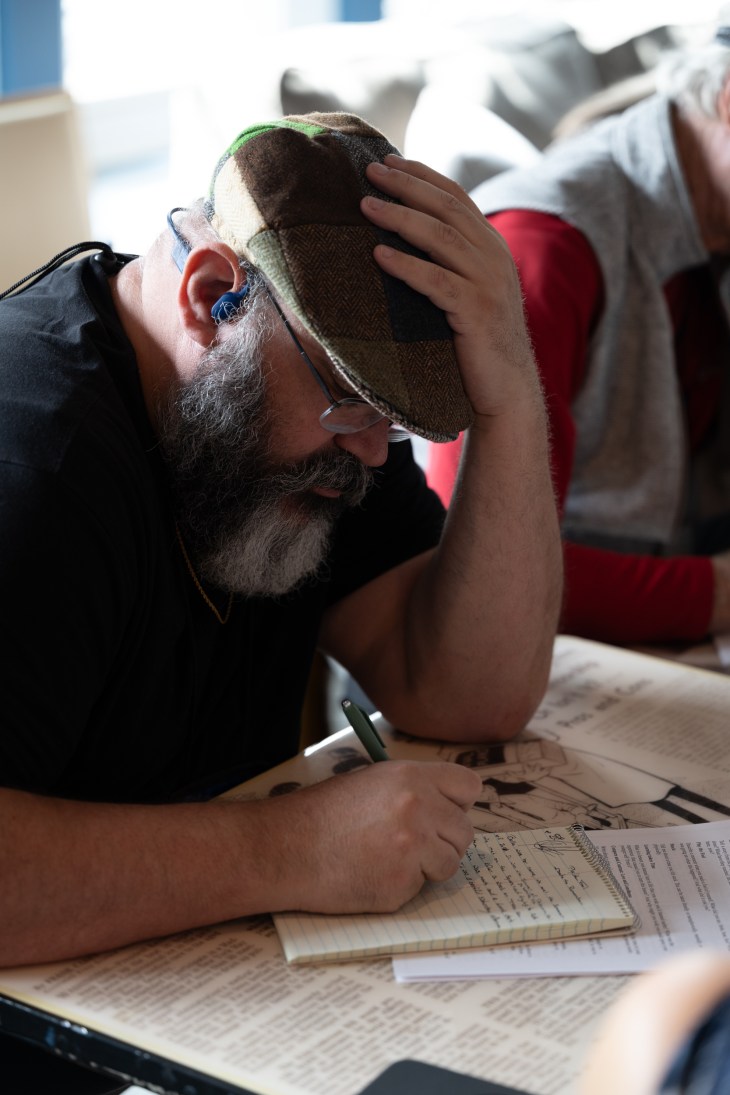 Man in black shirt and brown hat sits at a desk, writing on a notepad with his right hand while his left hand rests on his head.