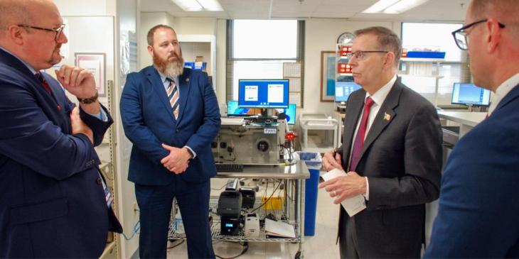 A group of four men wearing suits are standing in a laboratory setting, engaged in discussion. Monitors and lab equipment are visible in the background.